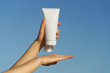 Female hands apply cream from white tube mockup on hand on blue sky background. The concept of...