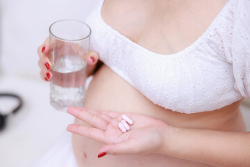 close up pregnant woman holding vitamin pill capsules and glass of water on hands.