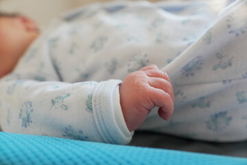 Newborn baby in a white onesie with blue designs, lying on a changing mat, hand visible, wrapped in...
