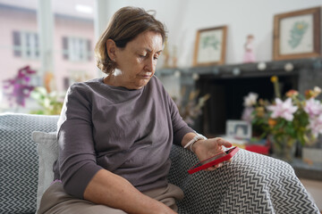 Elderly woman with short hair in a grey shirt sitting on a patterned couch, looking at her red...
