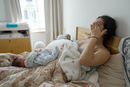 Mother showing exhaustion while sitting up in bed, covering her face with hands, and her newborn baby lying beside her, illustrating the fatigue and tender moments of postpartum recovery