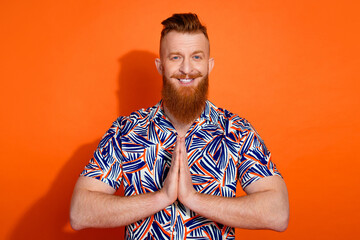 Photo of cheerful positive guy dressed print shirt arms together praying isolated orange color...
