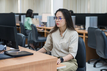 A woman sits at a desk in front of a computer. She is wearing glasses and is focused on her work....