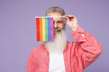Young elderly bearded gay man 50s years old wearing pink shirt casual clothes hold cover face eye with striped flag look camera isolated on plain purple background. Lifestyle LGBT June pride concept.