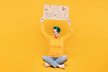 Full body young lesbian woman with dyed green hair wear hoody casual clothes hold cardboard with be yourself title text sit isolated on plain yellow orange background LGBT diversity lifestyle concept