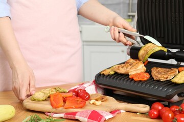 Woman cooking different products with electric grill at wooden table in kitchen, closeup