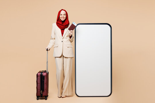 Traveler Asian Muslim woman wear red abaya hijab hold passport ticket bag big blank screen mobile cell phone isolated on plain beige background. Tourist travel, free time. Air flight trip UAE concept.