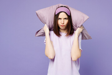Young sad mad woman wearing pyjamas jam sleep eye mask rest at home hold pillow cover ears from neighbours noise snoring isolated on plain pastel light purple background. Bad mood night nap concept.