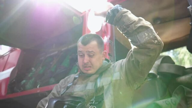 Male tired firefighter pouring water on his head from a bottle after extinguishing fire. Young fireman resting after emergency near a big red car. Concept of heroic profession. Close up Slow motion