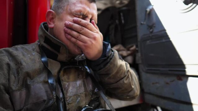 Young tired fireman wiping away the water on his face after emergency near fire engine. Male firefighter resting after extinguishing fire against background of big red car. Heroic profession concept