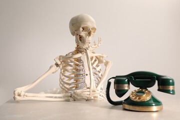 Waiting concept. Human skeleton at table with corded telephone against light grey background