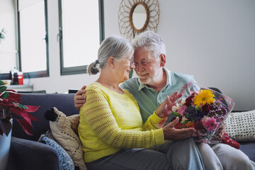 Senior man giving bouquet of flowers at his wife sitting on the sofa at home for anniversary or San Valentines’ day. Pensioners enjoying surprise together. In love people having fun.
