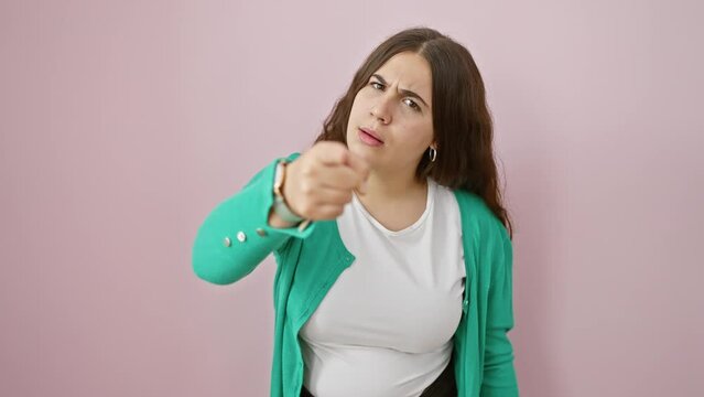 Frustrated, young hispanic woman accusingly points at you; furious and angry, standing isolated over a pink background, showing displeasure to the camera