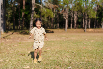 Asian girl in tan outfit running across grassy field in park, girl's smiling face reflects joy and...