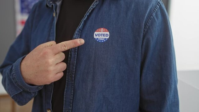 A man points to a 'voted today' sticker on his denim shirt, with an american flag blurred in the background.