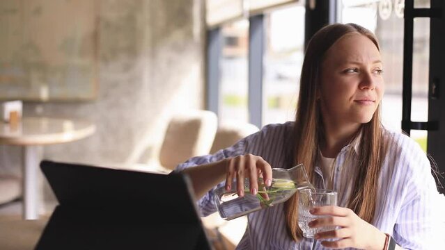 Woman pours water into a glass from a glass bottle. Blonde woman have a good time in cafe, she look happy and smiling. Girl wear stripped shirt. Woman drink water, look at side, happy, enjoy.