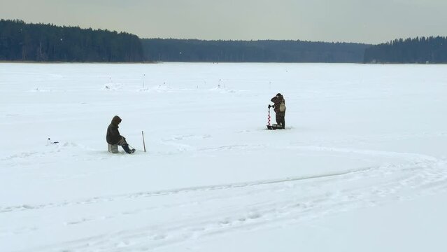 Fishermen on a winter fishing trip catch fish. Winter ice fishing. A winter fisherman is fishing on the pond. A man is sitting on the ice with a camping box, a drill and a thermos. Udmurtia. 4K	