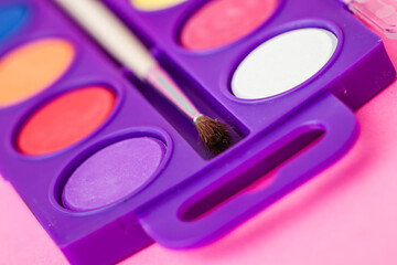 Close up of a watercolor paint palette with a paintbrush, vibrant colors on a pink background.