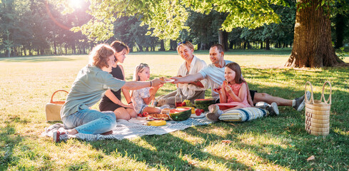 Big family sitting on the picnic blanket in city park during weekend sunny day. They smiling,...