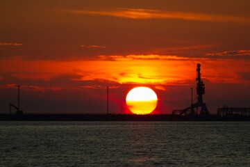 Fiery Sunset Over the Industrial Harbor. A Huge Orange Sun Descends Below the Horizon, Casting a Warm Glow on the Silhouettes of Cranes and Other Harbor Infrastructure.