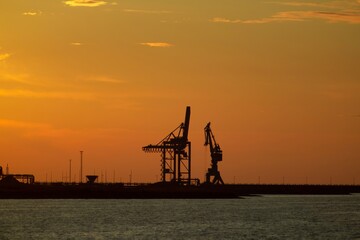 Obraz premium Silhouetted Industrial Cranes at Sunset, Creating a Dramatic and Peaceful Scene at the End of the Workday, Capturing the Beauty of Industrial Landscapes.