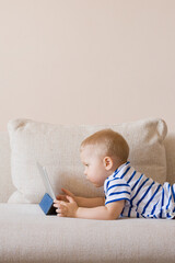 Adorable blond toddler boy sitting on the sofa and playing with tablet pc at home, indoors. Child with tablet computer. White cozy interior.