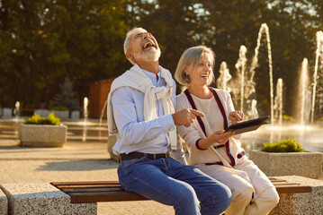 Happy senior couple finds joy and laughter while seated on a park bench, engrossed in technology...