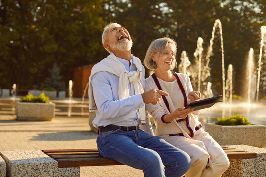Happy senior couple finds joy and laughter while seated on a park bench, engrossed in technology and communication. With a digital tablet computer in hand, they share laughter moments.