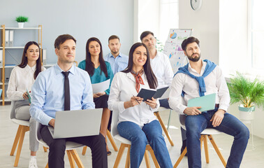 Group of smiling young business people sitting in a row on chairs and listening to colleague or their leadership at working place during business training or conference in meeting room.