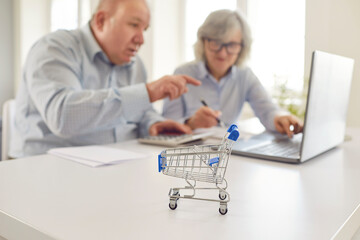 Close up shot of a toy shopping cart with an old couple, working on a laptop at home. Managing...