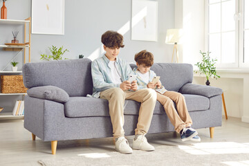 Two children brothers sitting on sofa at home holding mobile phones in hands playing online game or chatting in social media. The concept of kids technology and gadgets addiction problem.