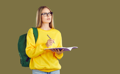 Serious thinkful blondy student girl in glasses and yellow sweatshirt with backpack makes notes in copybook on khaki background looking up on copy space. Education banner for advertisement concept.