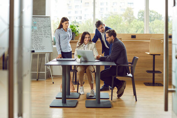 Business startup team using laptop during work meeting. Group of four young people gather around table with notebook computer in modern office workplace interior with whiteboard