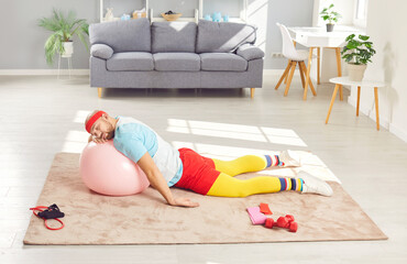 Funny lazy tired man takes a nap while working out. Weak weary young man in retro sports clothes and headband resting his head on yoga ball while sleeping on floor in living room after fitness workout
