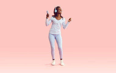 Full length portrait of a young joyful african american woman wearing casual clothes dancing and listening to music in headphones with mobile phone in hands isolated on a studio pink background.