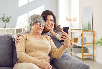 Elderly mother and adult daughter sitting together on a sofa at home, using a phone for a video...