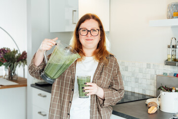 Smiling plus size young woman with just made glass of detox shake, green smoothie in the kitchen....
