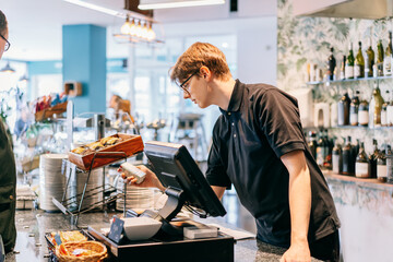 Young waiter serving customer at cash point in cafe. Man working with POS terminal. Cashier, barista checking for payment receipt. Hospitality, server and preparing a slip at the till in coffee shop.
