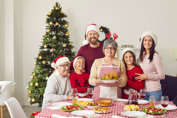 Portrait of large friendly multigenerational family at traditional Christmas dinner at home. Smiling older and younger family members in Santa hats gathered around older woman who is holding turkey.