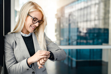 Smiling businesswoman checking the time on her watch while standing outside her office in modern...