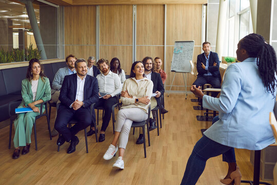 Diverse audience meeting with inspiring speaker at business seminar. Happy young and adult multiethnic businesspeople sitting in conference hall room listening to story by African American woman coach - Powered by Adobe