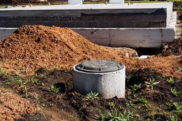 Construction in progress at site with concrete manhole cover and soil piles under sunny sky. Green...