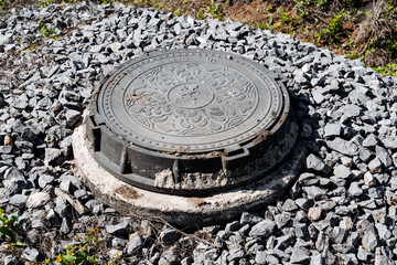 Ornate metal manhole cover surrounded by gravel stones in outdoor urban setting, showcasing decorative design. A functional utility cover providing access to underground infrastructure
