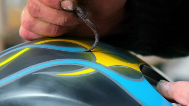 Close-up master hands,stained in paint,correcting airbrushing on motorcycle helmet using thin brush with long pile, applying frame to yellow blue fire flames ornament. Side view.Artist neat movements.