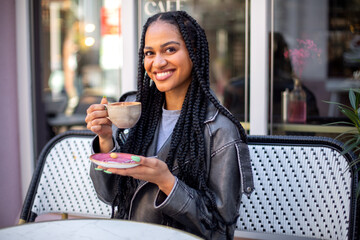 A happy young black female with afro braids is sitting outside the cafe with a cup of coffee