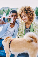Happy african american american young people together with dog dressed in casual wear smiling at front camera of modern smartphone device and making selfie photo for publishing in blog resting outdoor