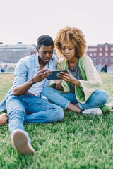 African american couple in love dressed in casual wear sitting on green grass and watching online broadcasting on smartphone using free 4G internet.Dark skinned friends searching information on tablet