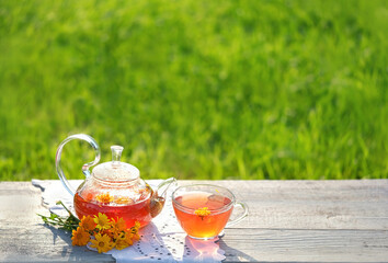 Glass Teapot and cup with herbal tea, fresh calendula flowers on wooden table in garden. Healing...