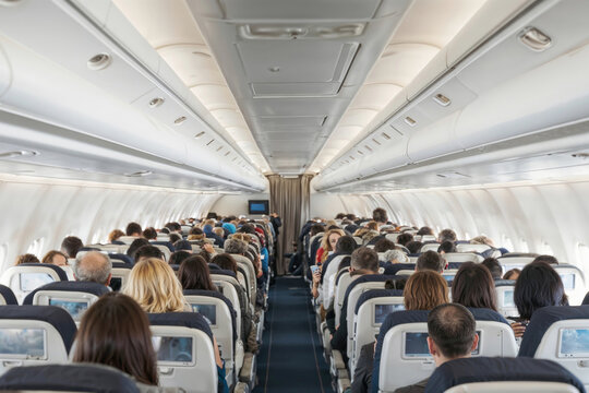 A crowded airplane with people sitting in rows. Inside Airplane Cabin. Walkway between rows of passenger seats in commercial airplane cabin