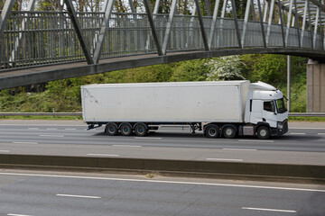 White, articulated lorry travelling on the the motorway  under the foot bridge in a sunny day with no other cars around.
